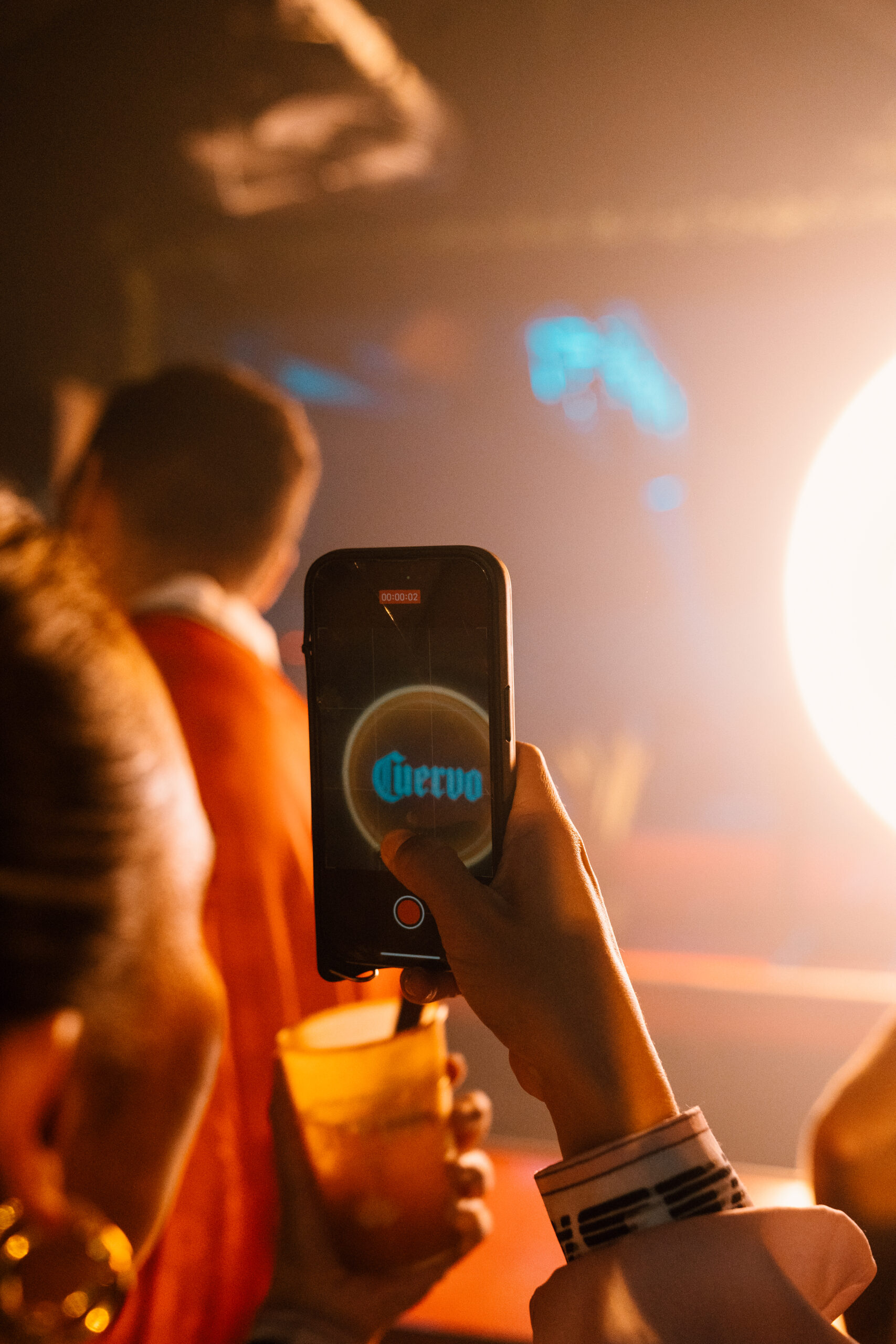 A sophisticated bar scene at Cuervo Cristalino Nights. In the foreground, a bartender expertly prepares a cocktail using a shaker. Bottles of Cuervo Cristalino tequila are prominently displayed on the bar, illuminated by warm lighting. In the background, guests can be seen socializing and enjoying drinks in a stylish and upscale atmosphere.