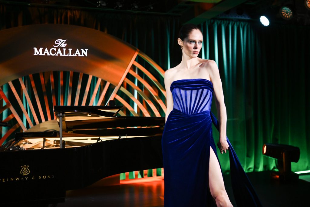 This image showcases a woman in a blue dress standing in front of a grand piano on a stage. The backdrop features the illuminated Macallan logo, creating a warm and inviting atmosphere. The backdrop itself is crafted from wood and brass, adding a touch of elegance and sophistication to the scene.